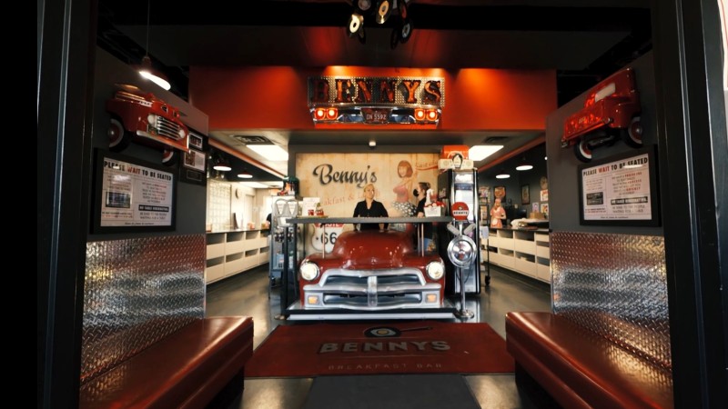 A retro-themed diner with a red car at the counter, Benny‘s sign above, and a welcoming atmosphere.