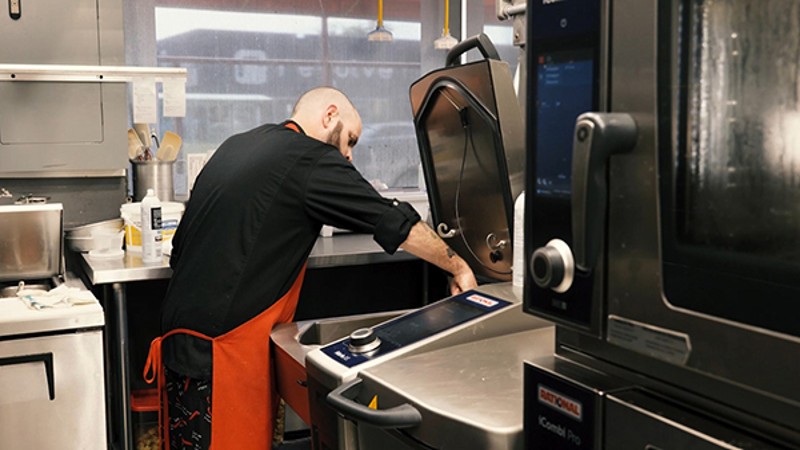 A chef in a black uniform works in the kitchen with cooking equipment visible, including a steam oven and a sous-vide machine.