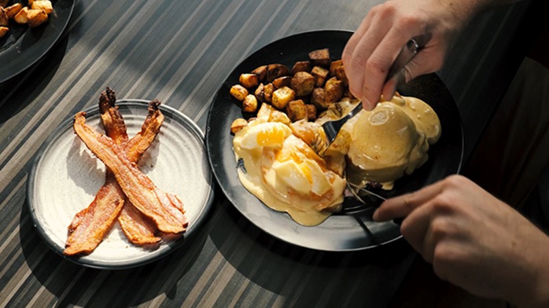 A close-up of a plate with bacon, potatoes, and eggs benedict, with someone using a fork and knife to cut the eggs.