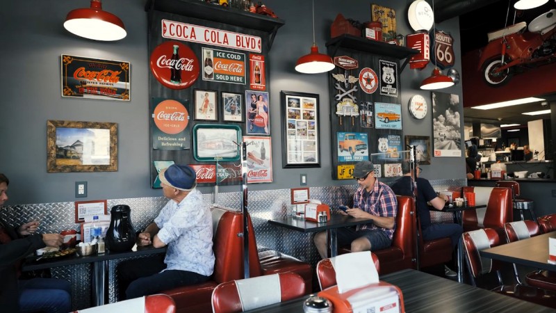 Retro diner interior with Coca-Cola and Route 66 signage, customers sitting at tables, and a vintage decor theme.