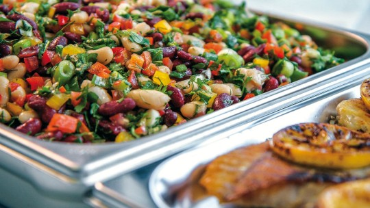 A close-up of a vibrant bean salad with peppers and herbs, alongside grilled fish and lemon slices in a catering tray.