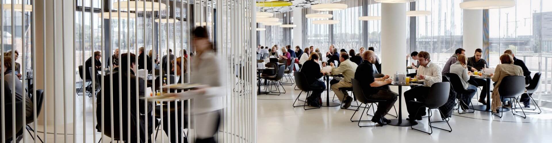 A modern cafeteria with circular ceiling panels and people seated at tables, while a staff member walks with a tray.