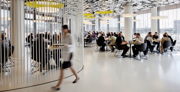 A modern cafeteria with circular ceiling panels and people seated at tables, while a staff member walks with a tray.