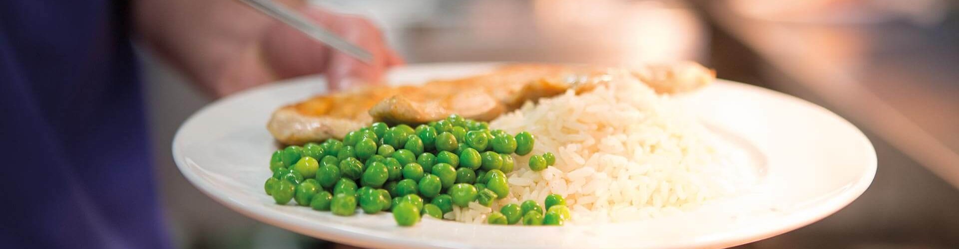 A hand serving a plate of food containing rice, green peas, and a piece of chicken in a busy kitchen setting.
