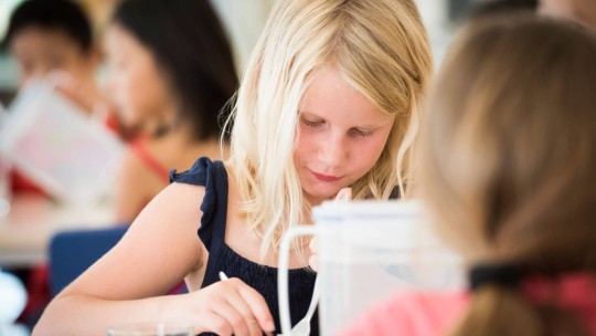 A young girl with blonde hair, focused on her meal in the school cafeteria, using a spoon.