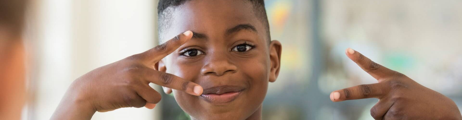 A young boy smiling and making a peace sign with his hands, dressed in a white t-shirt with a "cool" print.