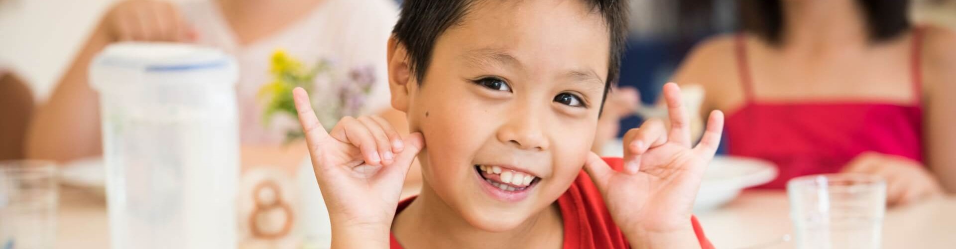 A young boy smiling and making hand gestures, with other children sitting at the table in the background.