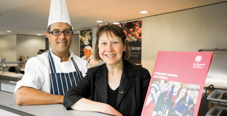 A smiling chef wearing a white chef hat and striped apron stands beside a woman with glasses.