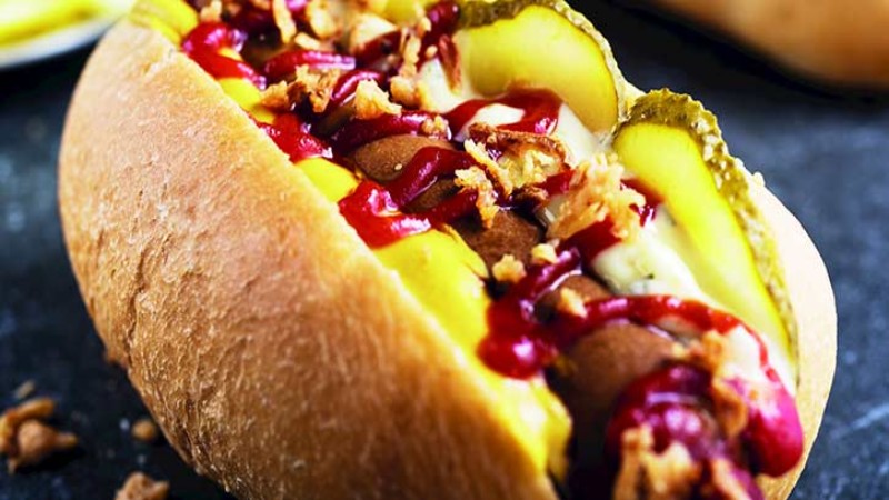 A delicious hot dog topped with mustard, ketchup, pickles, and crispy onions, served in a soft bun on a dark background.