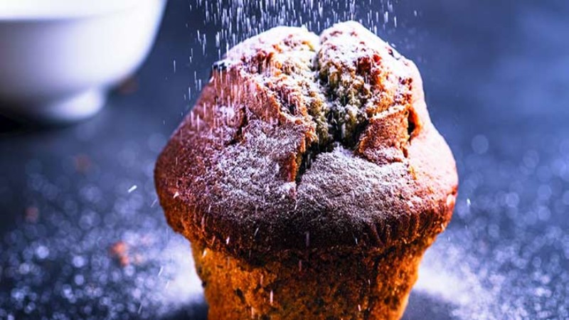 A freshly baked muffin dusted with powdered sugar, with a blurred bowl in the background, creating a cozy atmosphere.