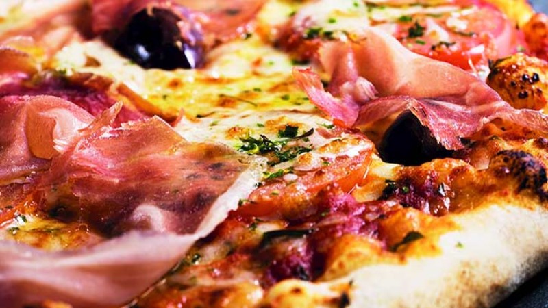 A close-up of a gourmet pizza topped with prosciutto, olives, and herbs, showcasing melted cheese and a crispy crust.