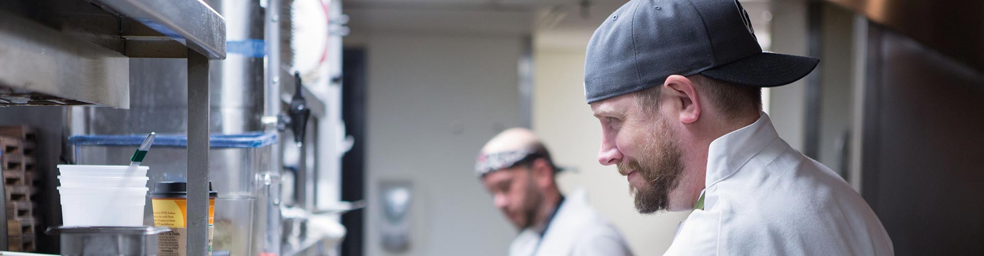 Two chefs in a busy kitchen, one wearing a cap, focused on meal preparation with various kitchen tools in the background.