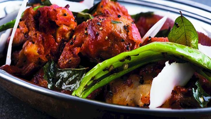 A flavorful dish of Chicken, featuring crispy chicken pieces garnished with green chili, curry leaves, and spices in a metal bowl.