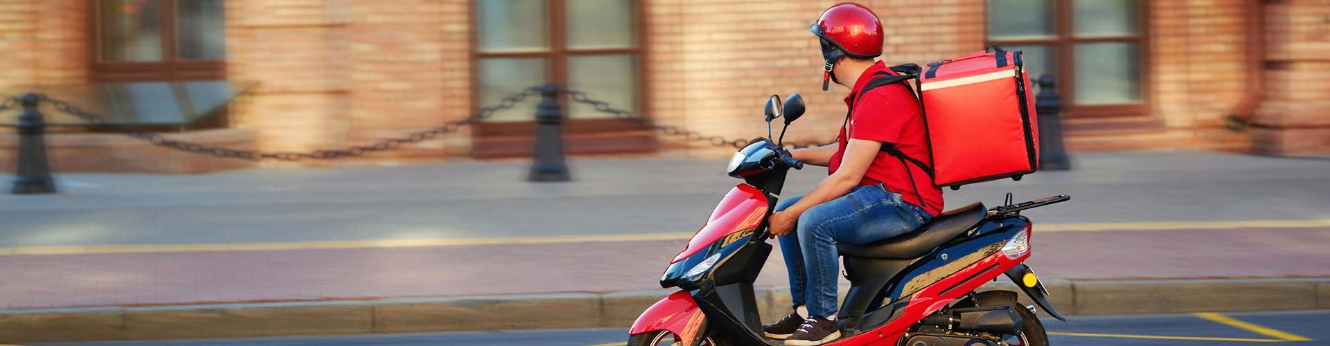 A delivery rider in a red helmet and shirt rides a scooter with a large red delivery bag on their back.