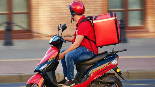 Un repartidor con casco y camiseta rojos conduce una scooter con una gran bolsa de entrega roja en la espalda.