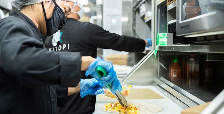 Un trabajador de cocina con guantes prepara un plato de tortilla en una cocina ocupada, con otro trabajador al fondo.
