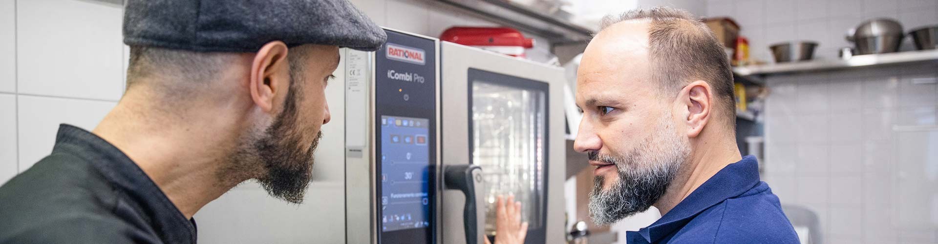 Two chefs discussing the operation of a Rational iCombi Pro oven, focusing on the control panel in a busy kitchen environment.