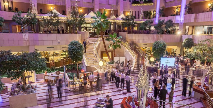 A beautifully lit hotel lobby with modern decor, staff serving guests, and a staircase leading to an upper level.