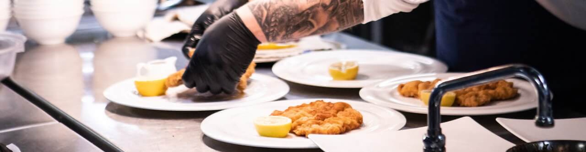 A chef in a ship galley plating a dish, wearing black gloves, with various ingredients and utensils in the background.