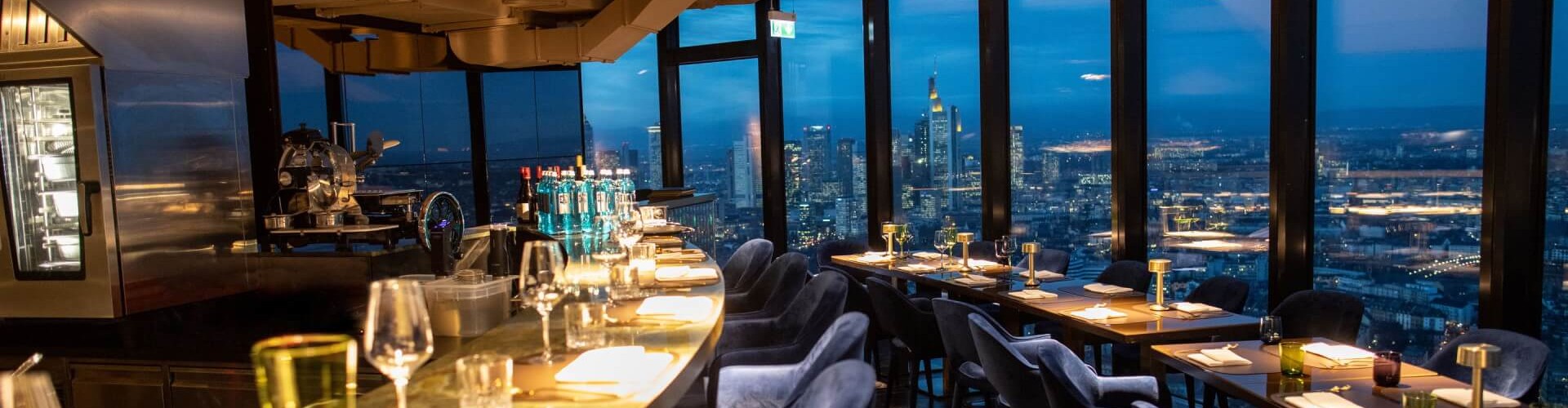 Un interior de restaurante moderno con una barra curva y mesas de comedor elegantes, que muestra vistas de la ciudad al anochecer, con copas de vino y elementos decorativos.