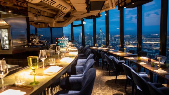 A modern restaurant interior with a curved bar and elegant dining tables, featuring city views at dusk, showcasing wine glasses and decorative items.
