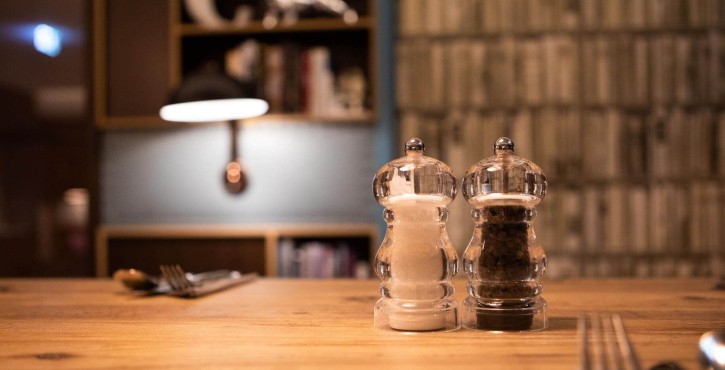 A close-up of a table setting featuring clear salt and pepper shakers on a wooden table, with utensils in the background.