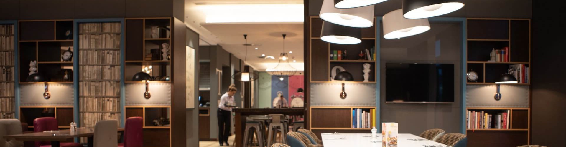 An elegant dining area with a long white table, stylish chairs, and modern pendant lights, featuring shelves with books.