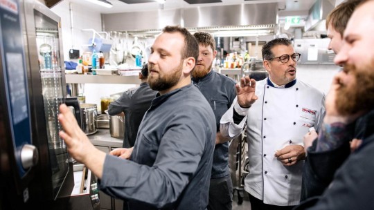 Eine Gruppe von Köchen versammelt sich um ein Rational iCombi Pro Gerät, während ein Koch die Funktionen erklärt und die anderen aufmerksam zuhören in einer belebten Küche.