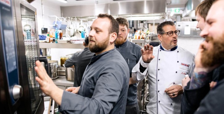 A group of chefs gathered around a Rational iCombi Pro appliance, with one chef explaining the features while others listen attentively in a busy kitchen.