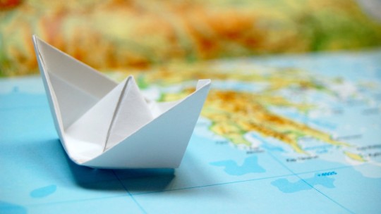 A white paper boat is placed on a blue map, symbolizing travel and adventure, with colorful land areas blurred in the background.