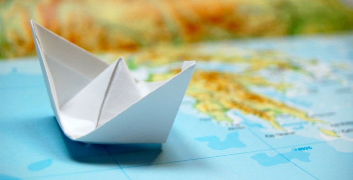 A white paper boat is placed on a blue map, symbolizing travel and adventure, with colorful land areas blurred in the background.