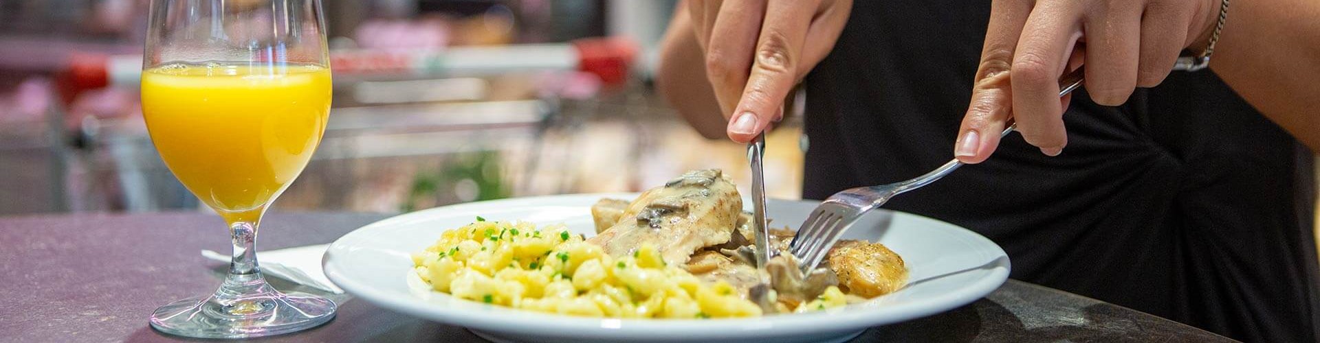 Eine Person hält eine Gabel und ein Messer, bereit, einen Teller mit Pasta und Hähnchen zu essen, begleitet von einem Glas Orangensaft.