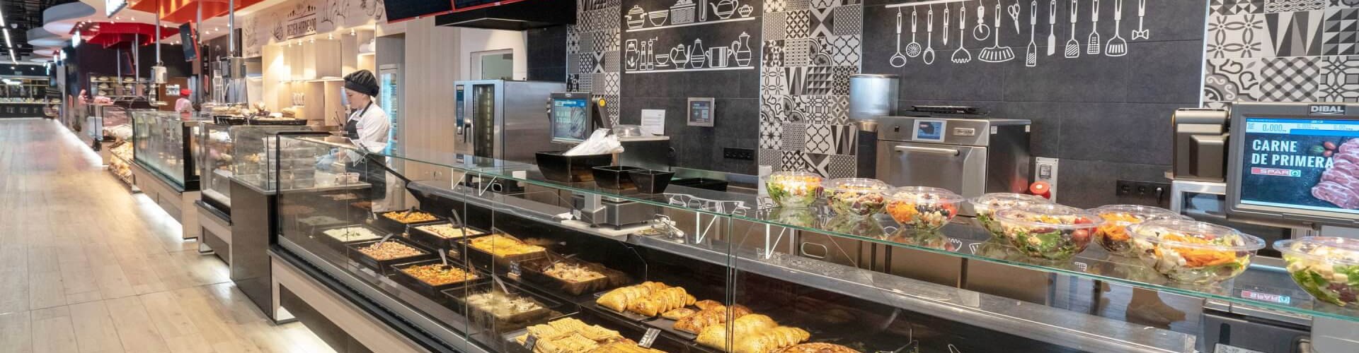 A view of a supermarket display counter with a variety of food options, including salads and baked goods, with a chef in a black hat preparing food in the background.