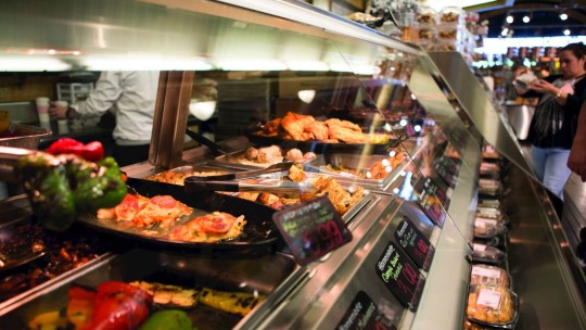 A selection of hot food displayed in a deli-style counter with various dishes, including grilled vegetables and meat, with prices visible.