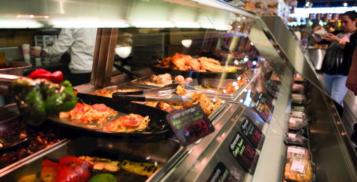 A selection of hot food displayed in a deli-style counter with various dishes, including grilled vegetables and meat, with prices visible.