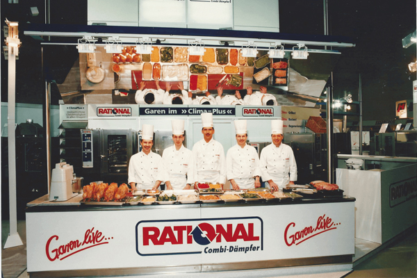 Historic photo of a RATIONAL trade fair booth with five chefs in white uniforms standing in front of a RATIONAL combi-steamer, showcasing various dishes and ingredients.