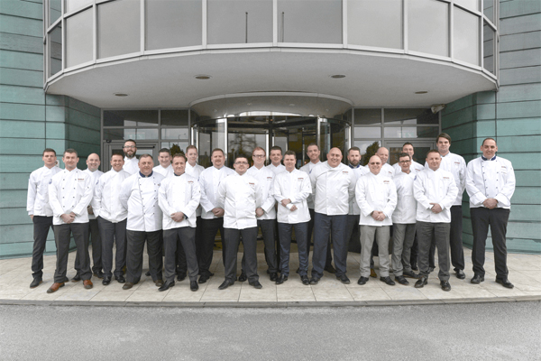 Several RATIONAL employees in white RATIONAL chef jackets are standing for a group photo outside a company building.