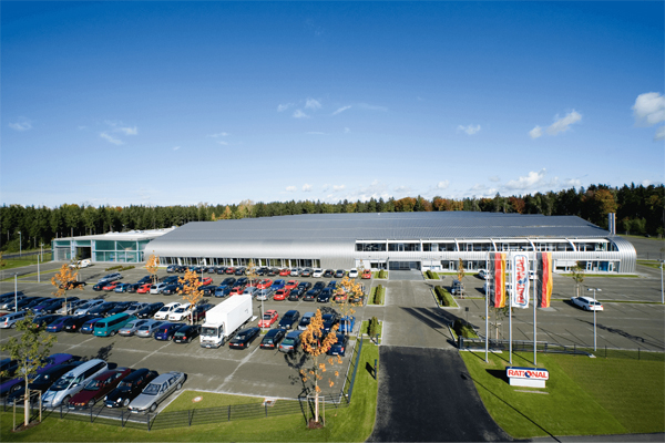 Outside view of RATIONAL AG’s Plant 3 with car park, company signage, and flags waving in sunny weather.