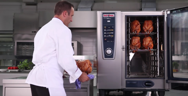 Chef in a white uniform holding a roasted chicken in front of a Rational combi-steamer.