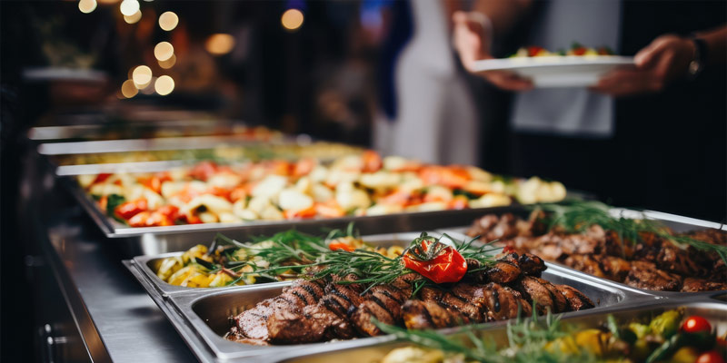 Buffet con platillos calientes variados, carne asada, vegetales y hierbas en evento de catering con autoservicio.