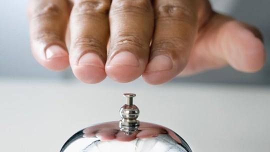 Close-up of a hand pressing a silver service bell on a white surface, typically used in reception or service areas.