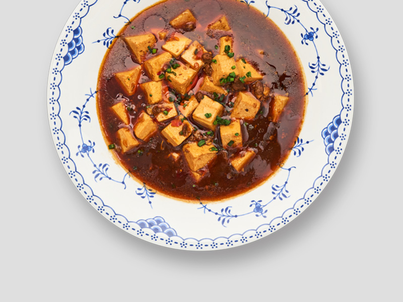 麻婆豆腐盛于蓝白花纹餐盘中，嫩豆腐与红油酱汁形成鲜明对比，点缀细香葱。