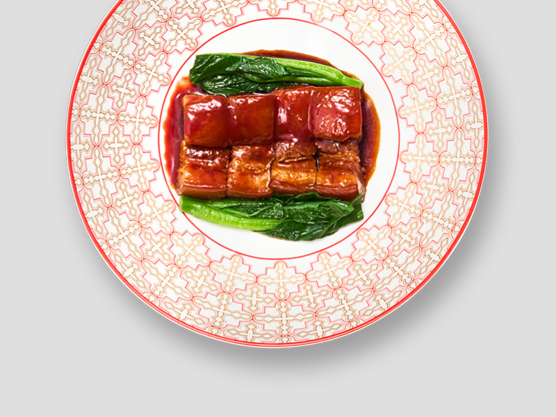 红烧肉块搭配绿叶蔬菜摆放在红纹图案餐盘中，色泽浓郁光亮，呈现经典中式焖煮风味。