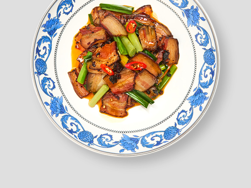 家常回锅肉搭配葱段与辣椒盛于蓝白花纹餐盘中，色泽红亮油润，呈现浓郁川味风格。