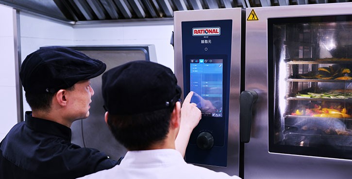 两位工作人员在操作RATIONAL 膳酷 混合蒸锅触控屏，旁侧设备内多层食材清晰可见。