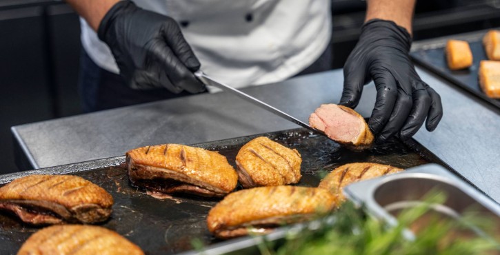 戴手套的工作人员在铁板上切开煎制肉块检查熟度，周围摆放多块金黄外皮的肉类。