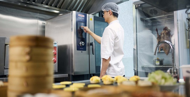 工作人员在厨房操作RATIONAL iCombi Pro 混合蒸锅触控屏，前景陈列多层蒸笼与点心。